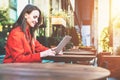 Side view.Young smiling attractive woman in orange coat is sitting outside in cafe at table and uses tablet computer. Royalty Free Stock Photo