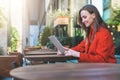 Side view.Young smiling attractive woman in orange coat is sitting outside in cafe at table and uses tablet computer. Royalty Free Stock Photo