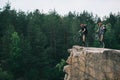 side view of young trial bikers with backpacks standing on back wheels on rocky cliff with blurred pine forest Royalty Free Stock Photo