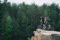 side view of young trial bikers standing on rocky cliff with blurred pine forest on background and looking Royalty Free Stock Photo