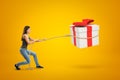 Side view of young woman standing with bent knees and pulling big gift box in air which she has lassoed, on yellow