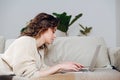 Side view of young woman working on a laptop, lying on a couch in her apartment