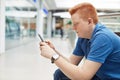 A sideways portrait of handsome redhead man with stylish hairdo dressed in blue shirt having a rest in shopping mall holding smart Royalty Free Stock Photo