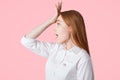 Sideways shot of beautiful female with long hair, keeps hand on forehead, regrets something, isolated over pink background. Lovely