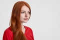 Sideways shot of beautiful red haired freckled woman with long hair, thinks about something, focused into distance, dressed in red