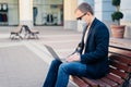 Sideways shot of busy man sits on wooden bench outdoor at station, waits for transport, uses modern laptop, connected to high