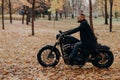 Sideways shot of confident male biker rides motorcycle, has adventure on two wheels in autumn park, wears stylish clothing,