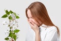 Sideways shot of displeased woman suffers from allergy, stands in front of branch with blossom, sneezes, feels hypersensitivity,