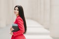 Sideways shot of elegant brunette lady with dark long straight hair, dressed in fashionable red suit, has manicure, carries diary