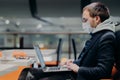 Sideways shot of freelancer keyboards on laptop computer, works on distance, wears protective medical mask during quarantine Royalty Free Stock Photo