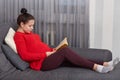 Sideways shot of pregnant young woman reads book, rests at home, sits at couch, dressed in red sweater, leggings and socks, enjoys