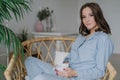 Sideways shot of pretty woman sits crossed legs in wicker chair, dressed in pyjamas, drinks hot coffee and latte, poses against co