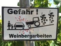 Sign in German that contains the words: Danger vineyard work. The sign in black and white shows vines, a tractor, a car and a moth