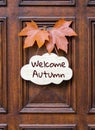 Sign with lettering words Welcome Autumn decorated with two orange maple leaves hanging on dark wooden entrance door.