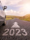 Sign 2023 on small asphalt country road and a side of a car and arrow. Travel and explore concept. Warm sunny day, sun flare. Royalty Free Stock Photo