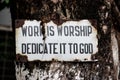 Sign with the words `work is worship dedicate it to god` ruined and hanging from a tree in Rishikesh