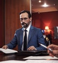 Serious bearded man in blue jacket, white shirt, tie filling documents sitting at table in office