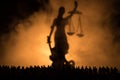 Silhouette Of Blurred Giant Lady Justice Statue With Sword And Scale Standing Behind Crowd At Night With Foggy Fire Background. At