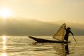 Silhouette Burmese fisherman wearing a hat standing at the back of a boat rowing with legs in the morning