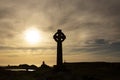 Silhouette of a Celtic cross with the evening sun. With a lighthouse, buildings and a cross in the background