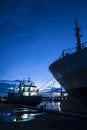 Silhouette fishing boat with many vessel and cargo ship docked at port against dark blue twilight sky background in vertical frame Royalty Free Stock Photo