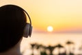 Silhouette of girl listening music in headphones standing on balcony and looking at sunset palm sea beach. Rear view of female. Royalty Free Stock Photo