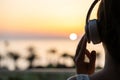 Silhouette of girl listening music in headphones standing on balcony and looking at sunset palm sea beach. Rear view of female. Royalty Free Stock Photo