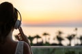 Silhouette of girl listening music in headphones standing on balcony and looking at sunset palm sea beach. Rear view of female. Royalty Free Stock Photo