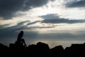 Silhouette of a girl looking towards the horizon, sitting on the seashore stones