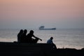 The silhouette of a group of unidentified people sitting on the embankment against the sea at sunset and a passing ship in the Royalty Free Stock Photo