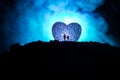 Silhouette of happy couple standing behind big shaped heart symbol on mountain at night. Big heart like moon glowing at foggy sky. Royalty Free Stock Photo