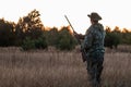 Silhouette of a hunter in a cowboy hat with a gun in his hands on a background of a beautiful sunset. The hunting period, the fall