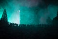 Silhouette of a large crowd of people in forest at night standing against a big arrow clock with toned light beams on foggy backgr