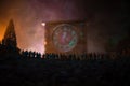 Silhouette of a large crowd of people in forest at night standing against a big arrow clock with toned light beams on foggy backgr