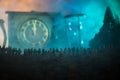 Silhouette of a large crowd of people in forest at night standing against a big arrow clock with toned light beams on foggy backgr