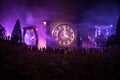 Silhouette of a large crowd of people in forest at night standing against a big arrow clock with toned light beams on foggy backgr