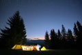 Silhouette of lonely hiker resting besides burning bonfire near illuminated tourist tents on camping site in dark