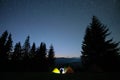 Silhouette of lonely hiker resting besides burning bonfire near illuminated tourist tents on camping site in dark