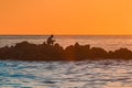The silhouette of a lonely man sitting in meditation on the rocks of a breakwater near the sea coast against the orange sunset
