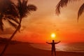 Silhouette of a man with his arms outstretched. Behind him is a beautiful sunset over the sea and the beach with palms
