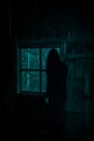 Silhouette of a man in a hood looking out the window in a scary dark attic of an old abandoned house in the darkness in Royalty Free Stock Photo