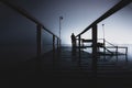 Silhouette of a man standing on a wooden dock in the dark staring at the emptiness of a lake with glowing lights on the opposite Royalty Free Stock Photo
