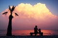 Silhouette mother and her toddler son sits on bench on an empty beach with lonely palm tree and looks at an amazing purple sunset