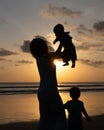 Silhouette of mother holding and lifting her infant baby boy high in the air on beach. Elder son standing nearby. Sunset time. Royalty Free Stock Photo