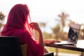 Silhouette of muslim, arab woman in red head scarf, hijab sitting at the table on terrace with laptop opposite sunset palm sea Royalty Free Stock Photo