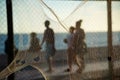 Silhouette of pedestrians, through a net, walking along the edge of a beach against the sunset