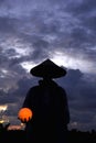 Silhouette of a person wearing traditional hat, carry ball lamp light in hand on blue dramatic sky cloud background. Royalty Free Stock Photo