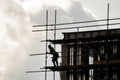 Silhouette scaffolder in PPE erecting framework of wooden planks and tall scaffolding poles high up on modern new building