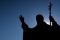 Silhouette of sculpture of pope John Paul The Second, with sun shining from behing him.