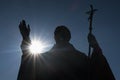 Silhouette of sculpture of pope John Paul The Second, with sun shining from behing his raised right arm.